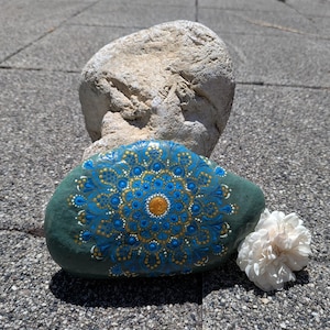 May include: A smooth, dark green river stone featuring a hand-painted mandala design in shades of bright blue, gold, and white. The intricate pattern consists of concentric circles of dots and teardrop shapes radiating from a central gold dot.