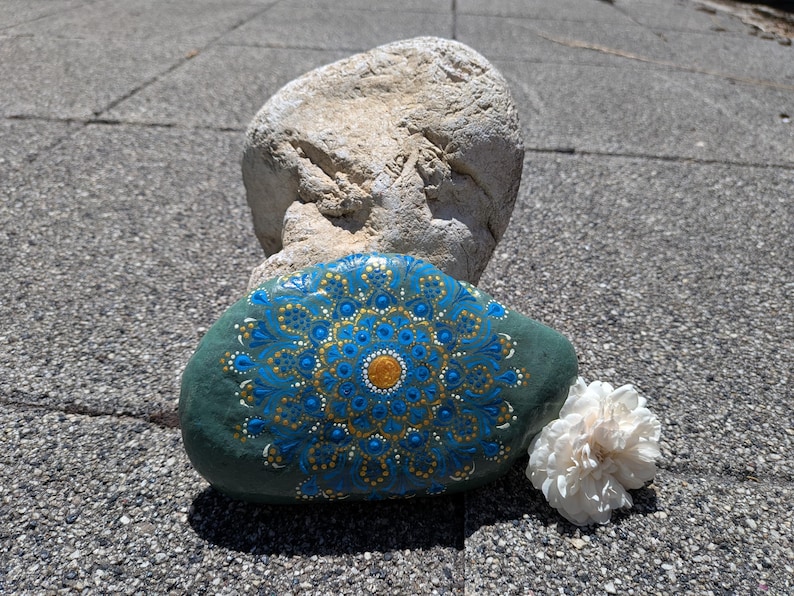 May include: A smooth, dark green river stone featuring a hand-painted mandala design in shades of bright blue, gold, and white. The intricate pattern consists of concentric circles of dots and teardrop shapes radiating from a central gold dot.