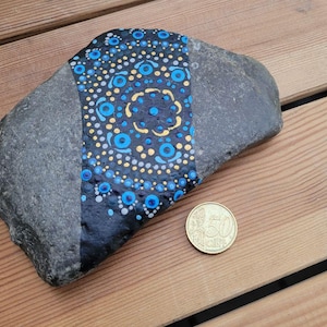 May include: A gray river stone featuring a hand-painted black band decorated with a symmetrical mandala pattern of blue, gold, and white dots. A 50-cent euro coin is placed next to the stone to provide a clear sense of scale for the object.