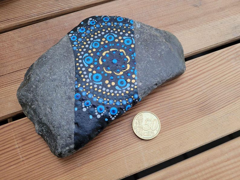May include: A gray river stone featuring a hand-painted black band decorated with a symmetrical mandala pattern of blue, gold, and white dots. A 50-cent euro coin is placed next to the stone to provide a clear sense of scale for the object.