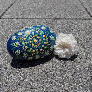 May include: A smooth, oval-shaped rock painted with a dark blue background and a detailed mandala pattern in yellow, white, and green dots. Beside the rock sits a single, ruffled white flower with many delicate, layered petals.