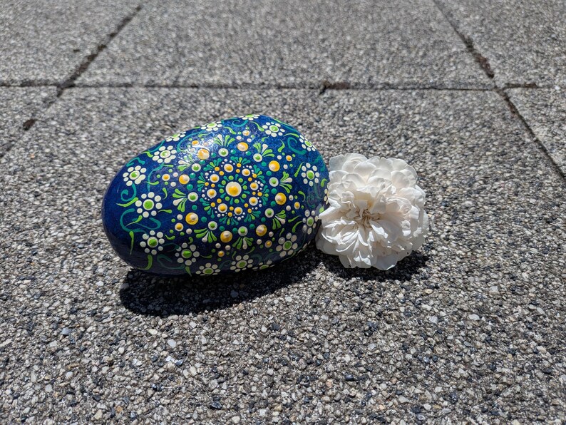 May include: A smooth, oval-shaped rock painted with a dark blue background and a detailed mandala pattern in yellow, white, and green dots. Beside the rock sits a single, ruffled white flower with many delicate, layered petals.