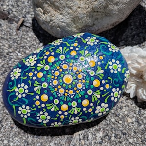 May include: A smooth, rounded stone painted with a vibrant blue base and a detailed mandala pattern. The design features intricate white, lime green, and yellow dots arranged in concentric circles and floral motifs across the surface.