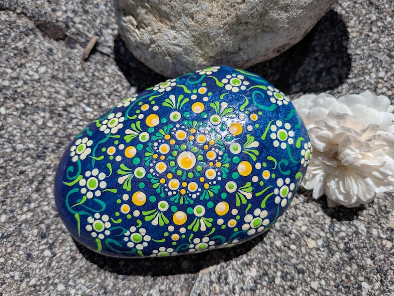 May include: A smooth, rounded stone painted with a vibrant blue base and a detailed mandala pattern. The design features intricate white, lime green, and yellow dots arranged in concentric circles and floral motifs across the surface.