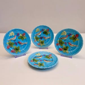 Może przedstawiać: Zestaw pięciu turkusowych ceramicznych talerzy. Każdy talerz przedstawia wzór ptaka z zielonymi liśćmi, czerwonymi jagodami i fioletowymi winogronami. Talerze są prezentowane na przezroczystych stojakach.