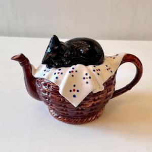 Op de afbeelding: Een keramische theepot met een mandweefpatroon, met een zwart kattenbeeldje op het deksel. De theepot heeft een bruine handgreep en tuit, en een witte doek met blauwe en rode stippen.