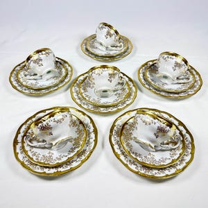Puede incluir: Un juego de seis tazas y platillos blancos con detalles florales dorados y ribetes dorados. Cada taza se asienta sobre un platillo a juego y un plato más pequeño. Las tazas tienen delicadas asas.