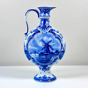May include: A blue and white ceramic pitcher with a handle and a flared spout. The pitcher features a hand-painted scene of windmills and water, with floral and geometric patterns. The pitcher stands on a pedestal base.