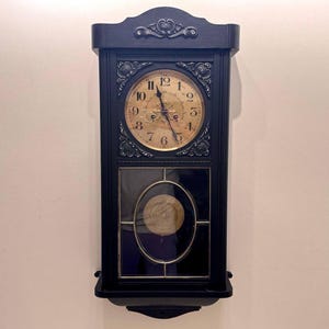 Könnte beinhalten: Eine schwarze antike Wanduhr mit goldfarbenem Zifferblatt und aufwendigen Verzierungen. Die Uhr hat ein Pendel, das durch ein Glasfenster sichtbar ist. Die Uhr hängt an einer weißen Wand.