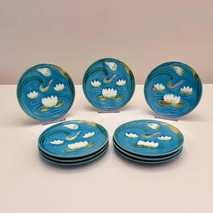 Może przedstawiać: Zestaw dekoracyjnych ceramicznych talerzy z motywem lilii wodnych. Talerze mają turkusowe tło z białymi liliami wodnymi, żółtymi akcentami i falistym wzorem. Talerze są okrągłe i ułożone w stos.