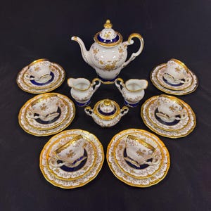 May include: A porcelain tea set featuring a teapot, a lidded sugar bowl, a creamer, and four cups with matching saucers. Each piece is white with deep blue bands, intricate gold floral patterns, and gold trim on the rims and handles.