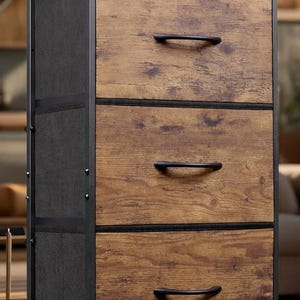 May include: A three-drawer chest with a rustic, industrial design. The drawers have a wood-grain appearance with black handles. The frame is black metal with mesh side panels. The chest sits on four short legs.