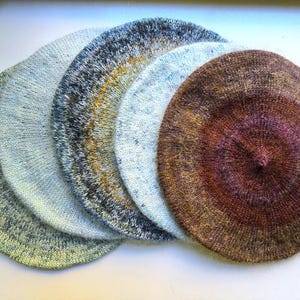 May include: Five circular knitted berets arranged in a fan shape. The berets feature varied color palettes including mottled green, light blue, dark grey with yellow accents, pale blue, and a gradient of deep brown, rust, and purple tones.