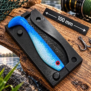 Puede incluir: Un molde rectangular negro para señuelos de pesca que contiene un cebo de plástico blando azul y blanco con un ojo rojo. Una barra de medición negra con texto blanco indica una longitud de 100 mm. El molde tiene pasadores de alineación y superficie texturizada.