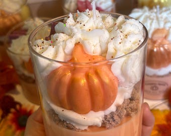 Op de afbeelding: Een heldere glazen kaars met een pompoenkruidengeur. De kaars heeft een kleine oranje pompoen, witte slagwas en een lichtbruine basis. Het etiket luidt "Pumpkin Spice 100% Soy Wax".
