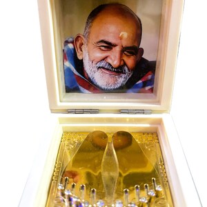 Puede incluir: Una caja blanca con bisagras con un retrato de un hombre sonriente en la sección superior. La sección inferior presenta una placa dorada con dos impresiones en forma de pie y elementos decorativos.