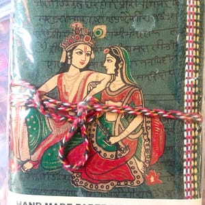 May include: A handmade paper notebook featuring an illustration of figures in traditional Indian dress. The green cover has a decorative border and is tied with a multi-coloured cord. The label reads "Hand Made Paper Note Book".