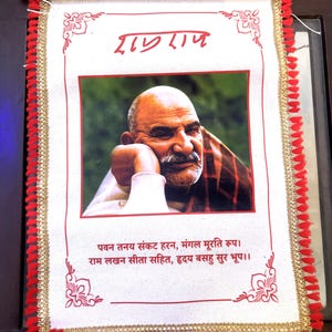 May include: A decorative wall hanging featuring a portrait of a man with a mustache. The image is framed with red trim and tassels, with text in a foreign language at the top and bottom. The background is a light color.