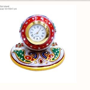 May include: A decorative quartz clock with a round, red and gold face, set in a matching stand. The clock face has Roman numerals and hands. The stand is white with colorful peacock designs. The text "Gol stand size 10 x 10 x 7 cm" is visible.