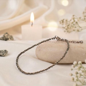 May include: A delicate pyrite bracelet with small, round beads. The bracelet is displayed on a smooth, light-colored stone, with a lit candle and soft fabric in the background. The bracelet has a silver-colored clasp.