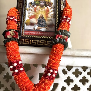 May include: A framed picture of a deity with an orange and red garland draped over it. The garland is made of folded ribbons and has white and red accents. The frame is dark brown with a gold pattern.