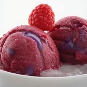 May include: Close-up of two scoops of berry-flavored ice cream in a white bowl, with a fresh raspberry on top. The ice cream is a deep red color with hints of purple. The ice cream is sitting on a bed of ice.