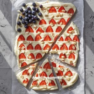 May include: A dessert bark with a white base, sliced strawberries, blueberries, and almond slivers arranged in a flag design. The bark is cut into irregular pieces, with the artist's name in the bottom right corner.