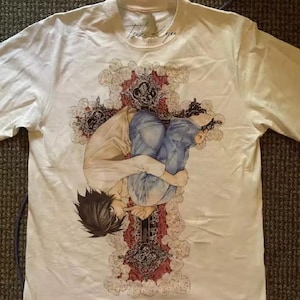 Puede incluir: Camiseta blanca con una ilustración detallada de anime. El diseño presenta un personaje en un patrón en forma de cruz, con cabello negro, una camisa clara y jeans azules. La camiseta incluye el texto "Tenka Kogen Studio".