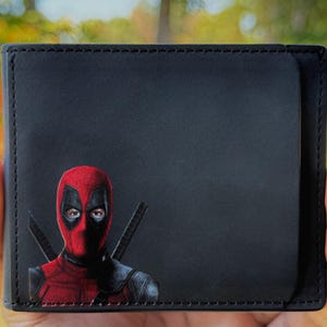 May include: Black bi-fold wallet featuring a Deadpool graphic. The graphic depicts Deadpool's face in a red mask with black accents and two swords. The wallet is made of a smooth, dark material.