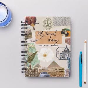 May include: A travel diary with a spiral binding and a collage cover with travel imagery and the text "My travel diary". A blue mug and a pen and pencil are next to the diary.