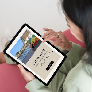 May include: A person holding a tablet displaying a book cover with the title "TRAVEL ALONE" and the subtitle "The guide to daring the world solo." The cover features a photo of a beach with colorful umbrellas.