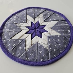 Op de afbeelding: Een ronde, gewatteerde onderzetter met een sterrenpatroon. De onderzetter heeft een centrale dieppaarse ster, omgeven door witte en blauwe stofsegmenten met patronen en een paarse rand. Een decoratief keukenartikel.