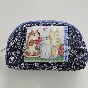 Op de afbeelding: Een marineblauwe, gewatteerde make-uptas met een bloemenpatroon. De tas heeft een rechthoekige patch met een cartoonillustratie van drie katten. De tas heeft een blauwe ritstrekker.