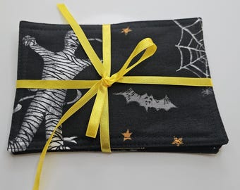 Pode incluir: Um conjunto de bases para copos pretas com tema de Halloween amarradas com uma fita amarela. As bases para copos apresentam uma múmia, morcegos, teias de aranha e estrelas em branco sobre um fundo preto. A fita está amarrada com um laço.