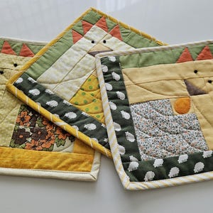 Peut inclure: Trois dessous de plat matelassés faits à la main avec un motif de poulet. Les dessous de plat présentent un motif patchwork avec des tissus jaunes, verts et orange. Les bords sont finis avec une bordure blanche. Les dessous de plat sont carrés et mesurent environ 20 cm sur 20 cm.