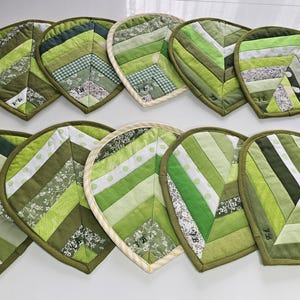 Puede incluir: Una colección de diez salvamanteles acolchados en forma de hoja en varios tonos de verde. Cada salvamanteles está hecho de retales de tela, creando un diseño de patchwork. Los bordes están acabados con un ribete verde oscuro.