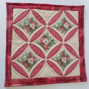Peut inclure: Un dessous de plat carré et matelassé avec un motif floral. Le dessous de plat présente un motif d'ovales roses et de carrés floraux, avec un fond crème. Les bords sont bordés d'un tissu rose plus foncé, et de petits boutons sont cousus aux intersections.