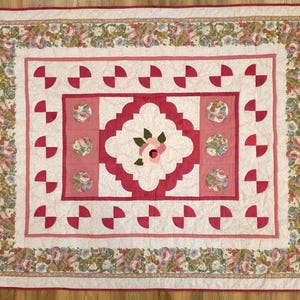 Pode incluir: Um edredom retangular com uma borda com padrão floral e um design central com uma flor rosa. O edredom tem um fundo branco com detalhes em rosa e vermelho, incluindo formas de semicírculo e medalhões florais.
