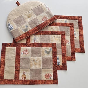May include: A quilted tea cozy and matching placemats. The set features a patchwork design with squares of beige, brown, and tan fabric. Each square has a cat illustration. The borders are a dark red-brown with a floral pattern.