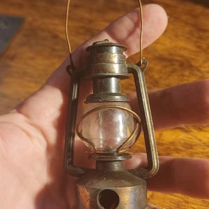 May include: A miniature antique-style lantern with a clear glass globe and a metal frame. The lantern has a small handle and a circular base. The metal has a tarnished, aged appearance. The lantern is held in a hand.