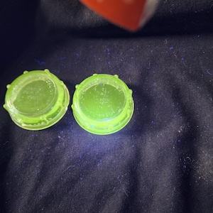 May include: Two circular, translucent neon green plastic caps with ribbed edges. The top surface of each cap features embossed text that reads "SEAL ON" and "CAP SEAL" with additional small, partially visible lettering in the center.