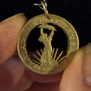 May include: A gold-colored pendant with a cutout design featuring a woman holding a leafy branch, surrounded by text. The pendant has a small loop at the top for a chain. The background is dark.