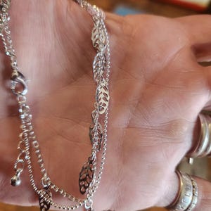 May include: A silver chain bracelet with a delicate leaf design. The bracelet is layered with two chains, one with a small charm at the end.