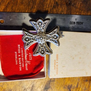 May include: A silver Christmas cross ornament with a textured design. The ornament is in its original box with a red velvet pouch. The box is labeled "1975 Christmas Cross - Limited Edition - Reed & Barton Sterling".