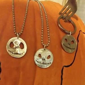 May include: Three Halloween-themed pendants and keychains. Two necklaces feature quarter coins with cut-out Jack Skellington faces, suspended on ball chains. A keychain with a similar coin design is also visible.
