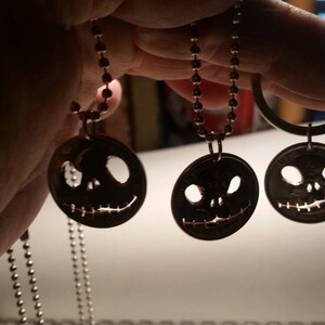 May include: Three dark metal pendants featuring Jack Skellington faces, each hanging from a silver ball chain. The round pendants have cut-out eyes and mouths, creating a spooky Halloween theme. The pendants are held up by a hand.