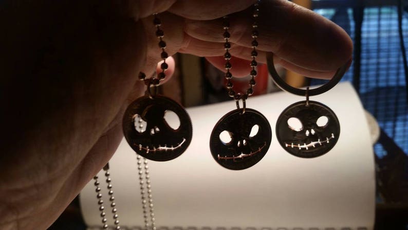 May include: Three dark metal pendants featuring Jack Skellington faces, each hanging from a silver ball chain. The round pendants have cut-out eyes and mouths, creating a spooky Halloween theme. The pendants are held up by a hand.