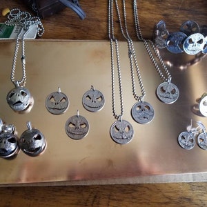 May include: A selection of silver-coloured jewellery with Jack Skellington designs. The set includes necklaces, pendants, a bracelet, earrings, and a pin. The jewellery is displayed on a reflective, gold-coloured surface.