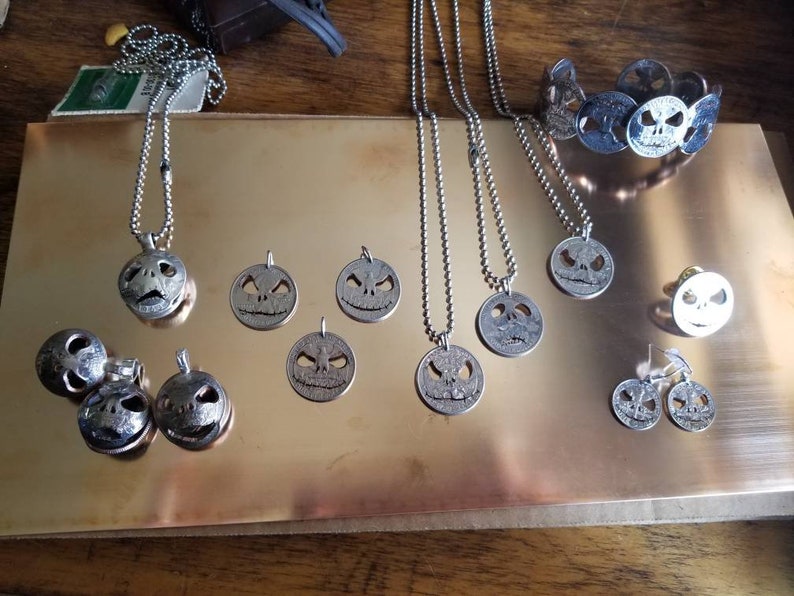May include: A selection of silver-coloured jewellery with Jack Skellington designs. The set includes necklaces, pendants, a bracelet, earrings, and a pin. The jewellery is displayed on a reflective, gold-coloured surface.