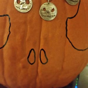 May include: Three silver coin pendants with carved faces, hanging on ball chains, and a keychain with a similar coin. The pendants and keychain are displayed on an orange pumpkin with black drawn features.